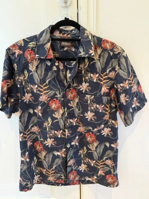 Oscar Jacobson kortärmad skjorta – Tropiskt blommönster – Strl S 🌺 - Snygg och stilren kortärmad skjorta från svenska Oscar Jacobson i storlek S. Skjortan har en mörkblå bas med ett elegant tropiskt blommönster i rött, orange, vitt och gröna toner – perfekt för sommar, semester eller en mer uppklädd casual look.  ✔️ Regular Fit ✔️ Kort ärm ✔️ Mjukt och bekvämt material ✔️ Stilfullt tropiskt mönster ✔️ Passar perfekt till chinos, jeans eller shorts ✔️ Mycket Fint skick  Från djur- och rökfritt hem.