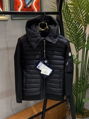 Svart pufferjacka från Moncler - Stilren svart pufferjacka från Moncler med quiltad front och huva. Jackan har släta ärmar med ribbade detaljer och en praktisk ficka med dragkedja på ena ärmen. Ikonisk Moncler-logga på ärmen och två framfickor med knappstängning. Perfekt för kalla vinterdagar.