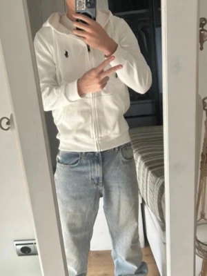 Vit Zip-hoodie - Snygg vit hoodie med dragkedja. Klassisk design med huva, dragsko och två fickor framtill. Liten broderad logga på bröstet. Perfekt för en clean och stilren look. Mjuk bomullskänsla och skön passform.