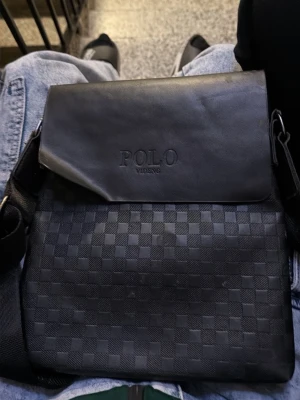 Svart axelväska från Polo Videng - Snygg svart axelväska från Polo Videng med lock och diskret rutmönster på framsidan. Väskan har justerbar axelrem, dragkedjeficka på baksidan och är tillverkad i konstläder. Perfekt för att bära dina viktigaste grejer med stil.