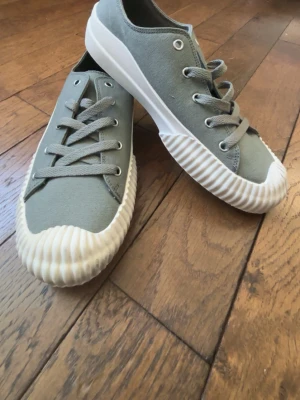 Grå sneakers med vit sula från XTI - Stilrena grå sneakers från XTI med lågt skaft och vit, räfflad gummisula. Skorna har ovandel i canvas och matchande grå skosnören. Helt nya och säljs eftersom dem var för små.