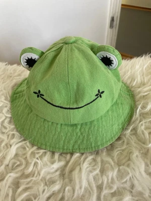 Groda Hatt För Vuxna Tonåringar, bucket hat med grodansikte - Söt grön groda har bucket hat i tyg med broderat grodansikte och utstickande ögon. Perfekt för dig som vill sticka ut med en lekfull och trendig accessoar. Hatten har en mjuk och bekväm passform och är tillverkad i bomullstyg.