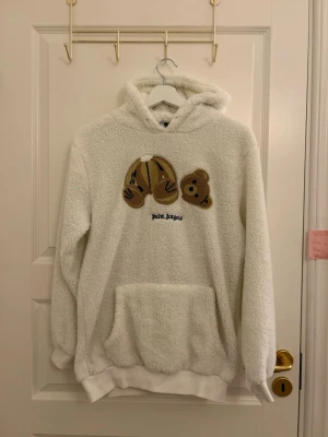 Vit teddyhoodie från Palm Angels - Mysig vit hoodie i teddy-material från Palm Angels med stor känguruficka och huva. På bröstet finns ett broderat motiv av en nallebjörn med avtagbart huvud och texten 'Palm Angels' under. Perfekt för dig som gillar streetwear och unika detaljer.
