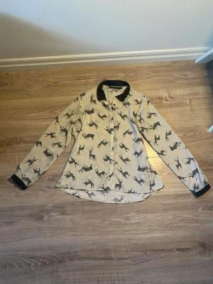 Beige skjorta med hästmönster - Stilren beige skjorta med svarta hästmönster och kontrasterande svarta detaljer på krage och ärmslut. Skjortan har klassisk passform och långa ärmar, perfekt för dig som gillar unika prints och vill sticka ut.