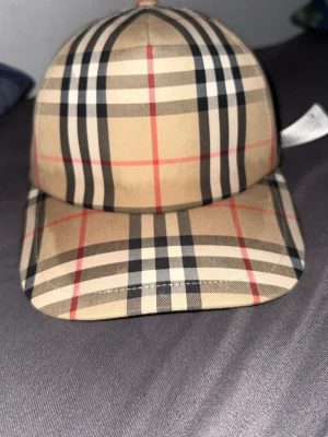 Beige keps med klassiskt rutmönster - Snygg beige keps från Burberry med det ikoniska rutmönstret i svart, vitt och rött. Kepsen har böjd skärm och justerbar passform. Perfekt accessoar för att lyfta din streetwear-look.