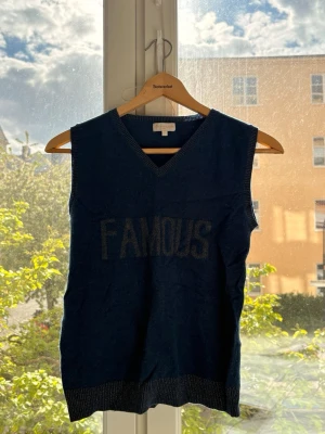 Mörkblå stickad väst från Bleu comme gris - Mörkblå stickad väst från Bleu comme gris Paris med v-ringning och texten 'FAMOUS' framtill. Västen har ribbade kanter i matchande färg och en klassisk, lite loose passform. Perfekt att styla över en skjorta eller t-shirt för en cool look.