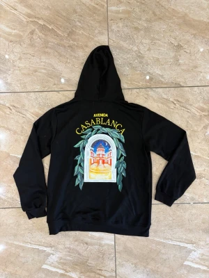 Svart hoodie från Casablanca - Svart hoodie från Casablanca med färgglad broderad grafik av en byggnad och texten 'Avenida Casablanca' på bröstet och ryggen. Hoodien har huva och ribbade muddar. Perfekt för dig som gillar streetwear och unika prints.