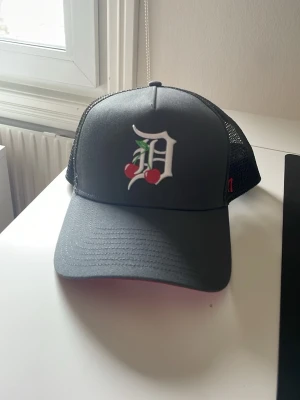 Svart keps med körsbär och Detroit tigers  - Snygg svart keps från 47 Brand med vit D-logga framtill, dekorerad med två röda körsbär och gröna blad. Kepsen har böjd skärm och mesh baktill för extra ventilation. Perfekt för en cool och avslappnad stil.