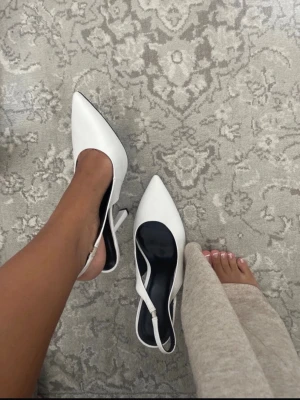 Vita slingback pumps med spetsig tå - Snygga vita pumps med spetsig tå och öppen häl, klassisk slingback-modell med smal klack. Skorna har en stilren design och justerbart spänne vid hälen. Perfekta för dig som gillar en elegant och trendig look.