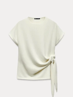 Vit topp med knytdetalj från Zara - Stilren vit topp från Zara med korta ärmar och rund halsringning. Toppen har en snygg knytdetalj i sidan som ger en trendig och modern look. Perfekt att matcha med både jeans och kjol. Mjukt och bekvämt material.