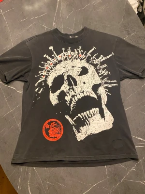 Svart Hellstar t-shirt med dödskalle -M - Cool svart t-shirt från Hellstar med stor vit dödskallegrafik och spikdetaljer på framsidan. Röd rund logga nertill. Klassisk passform och rund hals. Perfekt för dig som gillar edgy streetwear och vill sticka ut.