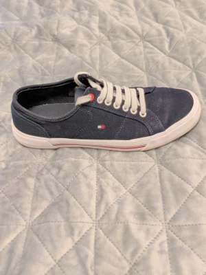 Mörkblå Tommy Hilfiger sneakers - Snygga mörkblå sneakers från Tommy Hilfiger med vita snören och vit sula. Skorna har diskret logga på sidan och klassisk låg modell. Ovandelen är i canvasmaterial och detaljer i rött och vitt ger en fräsch look.