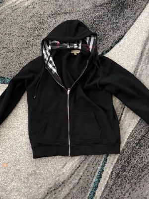 burberry zip - Svart hoodie med dragkedja från Burberry. Klassisk modell med två fickor fram och huva med ikoniskt rutigt foder i svart, vitt och rött. Perfekt för en stilren och avslappnad look. Materialet är mjukt och bekvämt, och huvan har dragsko.