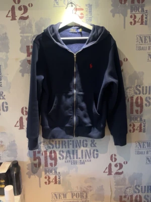 Marinblå hoodie från Polo Ralph Lauren - Snygg marinblå hoodie från Polo Ralph Lauren med dragkedja framtill och klassisk huva. Ikoniska röda loggan broderad på bröstet. Mjuk insida och ribbade muddar vid ärmslut och nederkant. Perfekt för en avslappnad och stilren look.