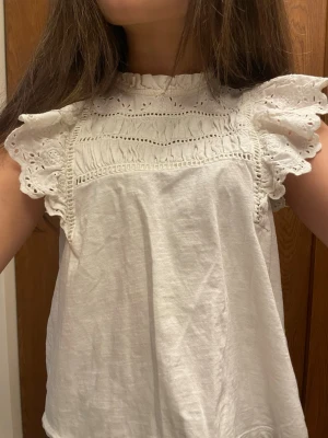 Vit blus med volangärmar och spets - Supersöt vit blus med broderade volangärmar och spetsdetaljer upptill. Blusen har rund halsringning med liten volangkant och är ärmlös med breda, romantiska volanger. Perfekt för dig som gillar en feminin och trendig stil. !🔥DENNA TRÖJA PASSAR ÄVEN PÅ EN S SOM EN LITE MER MAGTRÖJA MEN INTE ALS MYCKET🔥!