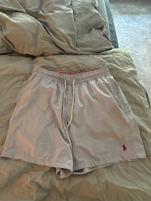 Grå shorts från Polo Ralph Lauren - Snygga grå shorts från Polo Ralph Lauren med elastisk midja och vit snörning. Insidan är röd och det klassiska Polo-loggan i rött syns på benet. Perfekta för sommardagar och chill vibes. Skönt material, lite tunnare ”fleece” material.