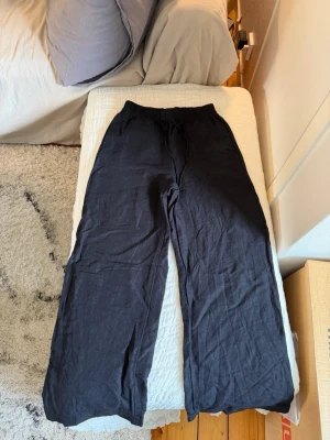 Svarta vida linnebyxor - Säljer ett par svarta linnebyxor med avslappnad passform och resår i midjan. Byxorna har raka, vida ben och är tillverkade i ett lätt och luftigt linnetyg. Perfekta för en chill och stilren look.