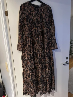 Blommig långklänning från Lindex - Långklänning från Lindex med svart botten och blommigt mönster i beige och bruna toner. Klänningen har lång ärm och rund halsringning, tillverkad i ett lätt och luftigt material. Perfekt för dig som gillar en bohemisk och avslappnad stil.