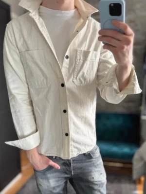 Vit overshirt med svarta knappar - Stilren vit skjorta med svarta knappar och två bröstfickor. Skjortan har lång ärm och klassisk krage, perfekt för en clean och enkel look. Tillverkad i bomull för en skön känsla och avslappnad passform.