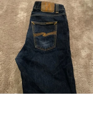 Mörkblå jeans från Nudie Jeans - Säljer ett par mörkblå jeans från Nudie Jeans med klassisk femficksdesign och snygga kontrastsömmar på bakfickorna. Jeansen har en rak passform och är tillverkade i denim med tydliga detaljer och läderpatch bak i midjan.