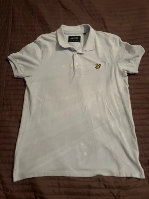 Ljusblå pikétröja från Lyle & Scott - Snygg ljusblå pikétröja från Lyle & Scott med klassisk krage och knappar framtill. Tröjan har korta ärmar och den ikoniska gula loggan broderad på bröstet. Perfekt för en stilren och avslappnad look.