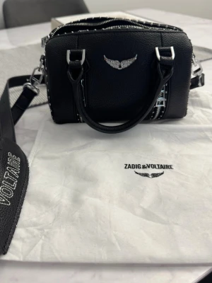 Svart handväska Zadig & Voltaire - Cool svart handväska från Zadig & Voltaire i strukturerat läder med silverfärgade metalldetaljer och nitar längs kanterna. Väskan har dubbla handtag, avtagbar axelrem och ikonisk vinglogga framtill. Perfekt för dig som gillar edgy och stilren design.