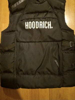 Svart dunväst från Hoodrich - Svart dunväst från Hoodrich med stor vit logga på bröstet. Västen har justerbara spännen på sidorna och en stor ficka framtill. Puffig design och hög krage ger en streetwear-look. Perfekt för lager-på-lager-stil under kyliga dagar.