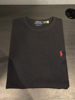 Svart t-shirt från Polo Ralph Lauren - Svart t-shirt från Polo Ralph Lauren med klassisk passform och rund hals. T-shirten har en liten röd broderad logga på bröstet och är tillverkad i mjuk bomull. Perfekt för en stilren och enkel look.