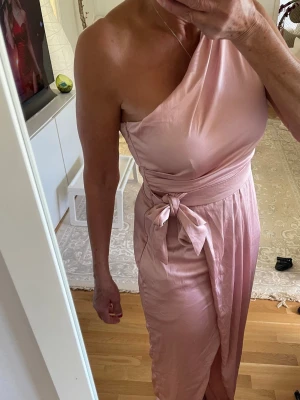 Rosa oneshoulder långklänning med knyt - Elegant rosa långklänning i satinliknande material med oneshoulder-design och bredt midjeband som knyts i sidan. Klänningen har en slits framtill och mjukt fallande tyg som ger en lyxig känsla.en dragkedja på sida av klänningen 