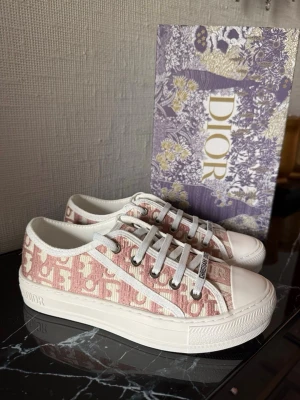 Dior skor rosa - Snygga Dior sneakers i rosa och vit canvas med klassiskt Dior-monogram. Skorna har vita snören, gummisula med logga och detaljer i vitt. Perfekt för dig som vill sticka ut med exklusiva och trendiga sneakers från ett lyxigt märke.