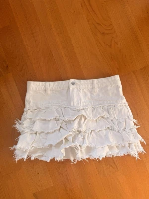 Vit volangkjol från Monki - Supersöt vit kortkjol från Monki med flera lager volanger och råa kanter för en trendig look. Kjolen har bälteshällor och knapp framtill. Perfekt för dig som vill sticka ut med en lekfull och edgy stil. Strl 36