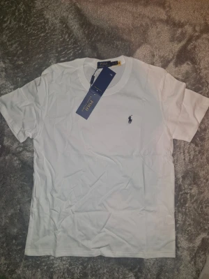 Vit t-shirt från Polo Ralph Lauren - Klassisk vit t-shirt från Polo Ralph Lauren med broderad marinblå logga på bröstet. T-shirten har rund halsringning och korta ärmar. Tillverkad i mjuk bomull för en skön känsla.
