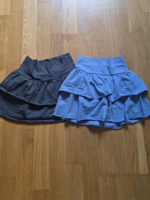 Två volangkjolar i blått och svart - Säljer två korta kjolar med volang i blått och svart. Kjolarna har bred midjeresår och dubbla volanglager som ger en söt och lekfull look. Perfekta för dig som gillar en avslappnad men trendig stil.