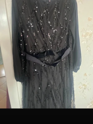 Svart långklänning med paljetter - Elegant svart långklänning med transparenta långärmar och glittriga paljettdetaljer över hela klänningen. Klänningen har ett band i midjan och är perfekt för speciella tillfällen. Materialet känns lätt och festligt.