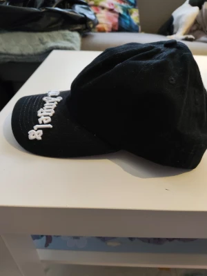 Svart Palm Angels keps - Säljer en svart keps från Palm Angels med böjd skärm och vit broderad logotyp framtill. Klassisk modell med justerbar rem baktill. Perfekt för dig som vill ha en cool och stilren accessoar med streetwear-vibe.