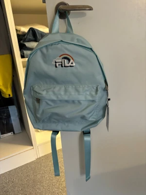 Ljusblå ryggsäck från FILA - Snygg ljusblå ryggsäck från FILA med regnbågsbroderi och logga på framsidan. Tillverkad i slitstark polyester med justerbara axelband och en praktisk ytterficka med dragkedja. 