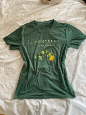 Grön t-shirt med citrontryck strl s-m - Snygg grön t-shirt med texten 'Limoncello' och ett citrontryck framtill. Modellen är klassisk med rund hals och korta ärmar. Perfekt för dig som gillar fräscha prints och vill sticka ut lite extra.                                                              Storlek eller märke står inte men skulle säga att den passar som en lösare s eller tajtare m