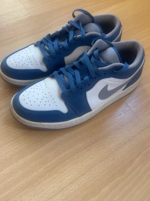 Nike Air Jordan 1 Low blå/vit - Snygga Nike Air Jordan 1 Low sneakers i blått och vitt med grå swoosh. Skorna har klassisk låg siluett, perforerad tåbox och vadderad plös. Ovandelen är i läder och textil, med blå snörning och grå insida. Perfekta för dig som gillar streetwear och sportig stil.