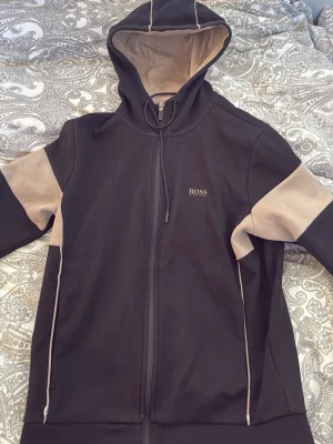 Svart och beige set från BOSS - Snyggt svart och beige set från BOSS med dragkedja på tröjan och huva. Setet består av en hoodie och matchande byxor med beigea detaljer på ärmar och ben. BOSS-logga på både tröja och byxa. Perfekt för en avslappnad och trendig stil.