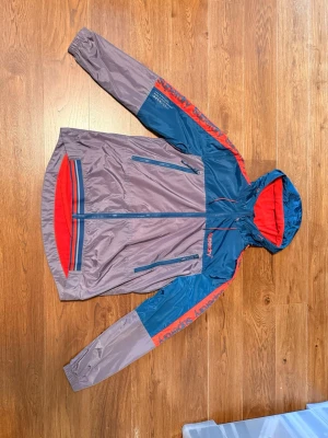 Vindjacka från superdry i blått och grått - Snygg vindjacka från superdry i blått och grått med röda detaljer. Jackan har huva, dragkedja framtill, två sidofickor med dragkedja och röd meshfoder på insidan. Logga på bröstet och längs ärmarna. Perfekt för blåsiga dagar.