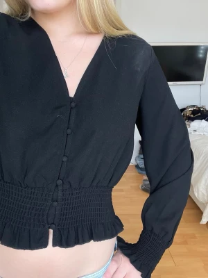Svart blus med smock och knappar - Trendig svart blus med smockad midja och ärmslut, dekorativa knappar framtill och v-ringning. Blusen har långa ärmar och volangdetalj nedtill. 