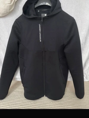 Svart vindjacka från Under Armour - Säljer en svart vindjacka från Under Armour med huva och reflekterande detaljer fram och bak. Jackan har dragkedja hela vägen och är tillverkad i ett lätt syntetmaterial. Perfekt för dig som vill ha en sportig look och gillar funktionella plagg. Helt oanvänd och fräsch!