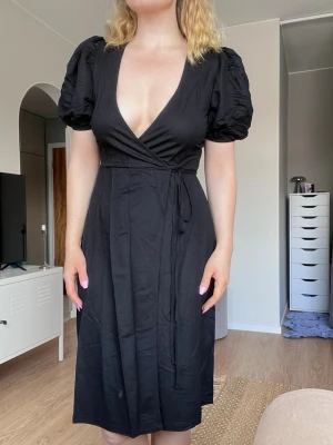 Svart omlottklänning med puffärm från Cubus, storlek XS (nyskick) - Svart omlottklänning med djup v-ringning och puffiga kortärmar. Knytband i midjan och går ner till knäna. Använd en gång, superfint skick!
