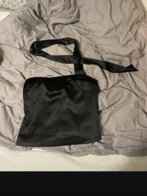 Svart satin tubtopp med knytband - Snygg svart tubtopp i satin med breda knytband som kan knytas runt halsen. Toppen har en glansig finish och är axelbandslös, perfekt för en trendig look.