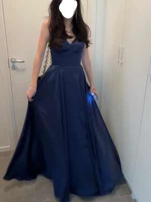 Marinblå långklänning med slits - Elegant marinblå långklänning med v-ringad urringning och breda axelband. Klänningen har en snygg slits framtill som ger en modern touch och ett svepande fall. Perfekt för bal eller festliga tillfällen. Materialet har en lätt glansig finish.