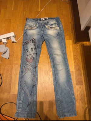 Blå jeans med Betty Boop-tryck - Unika blå jeans från La visione med coolt Betty Boop-motiv på ena benet och mönstrade detaljer vid fickorna. Jeansen har raka ben och slitna partier för en trendig look. Perfekta för dig som vill sticka ut med din stil.