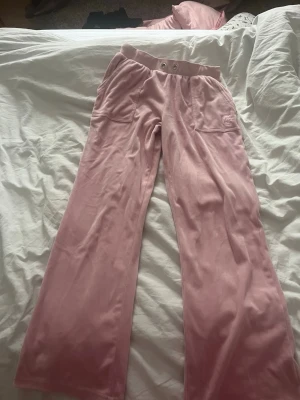 Rosa mjukisbyxor Juicy Couture - Säljer ett par klassiska rosa mjukisbyxor från Juicy Couture i mjuk velour. Byxorna har raka ben, bred resår i midjan med två knappar och fickor fram. Juicy Couture-logga broderad på ena fickan. Perfekta för en chill och trendig look.