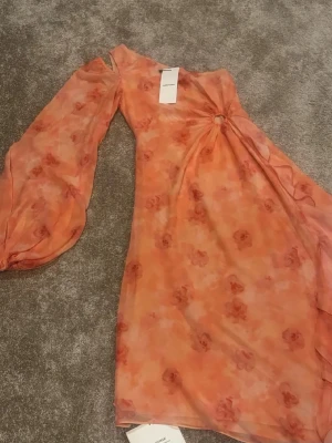 Adoore klänning peach strl 34 - Säljer en peachfärgad one shoulder klänning från Adoore med blommigt mönster och puffärm. Klänningen har en snygg ringdetalj framtill och är gjord i ett lätt, transparent material som känns som polyester. Perfekt för sommarens alla tillfällen! Helt ny med tags! Nypris: 1695kr