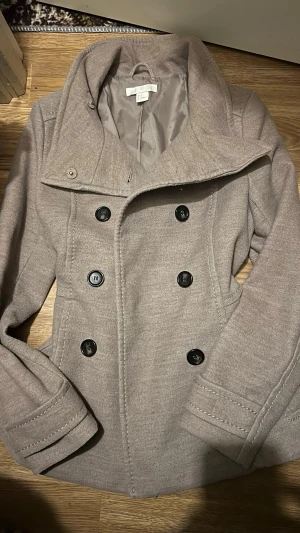 Peacoat - Snygg beige rock från H&M med dubbelknäppning och stora svarta knappar. Rocken har hög krage, figurnära passform och två fickor framtill. Tillverkad i ett mjukt tyg som ger en stilren look. Perfekt för dig som vill ha en klassisk men modern stil.