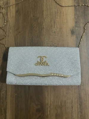 Glittrig silver kuvertväska från Chanel - Snygg kuvertväska från Chanel i glittrigt silver med guldfärgade detaljer och kedja. Väskan har ett elegant CC-logo framtill och en vågig guldfärgad dekor på locket. Perfekt för att lyfta din outfit med lite extra bling.